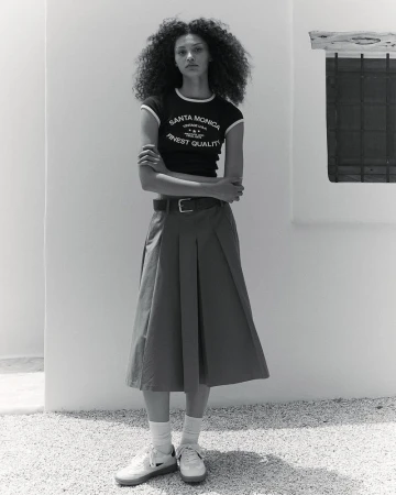 Full shot of a young, slim adult female with curly hair in a dark tee and long pleated skirt, posing outdoors in a minimalist style.