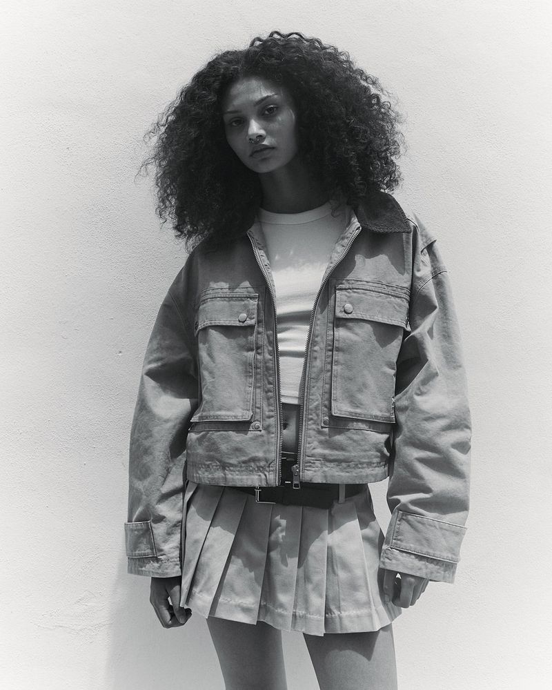 Model Enya Davis wearing a cropped utility jacket and pleated mini skirt in a black and white portrait against a light backdrop.