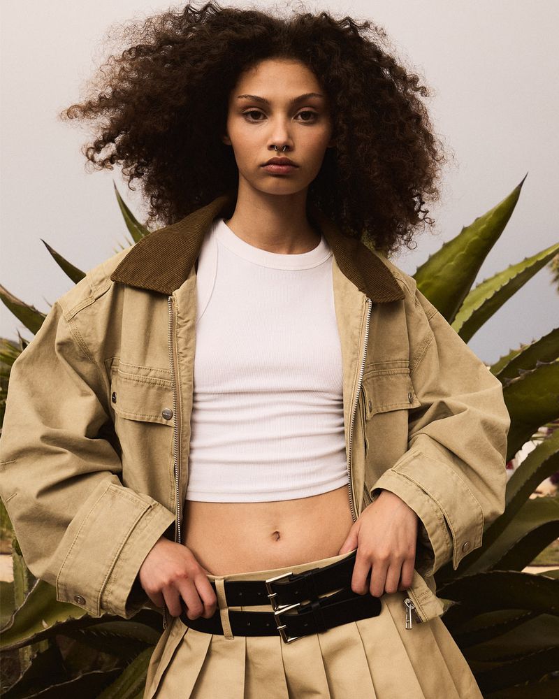Portrait of a young adult female with curly brown hair wearing a khaki jacket and pleated skirt, standing outdoors against a leafy background.