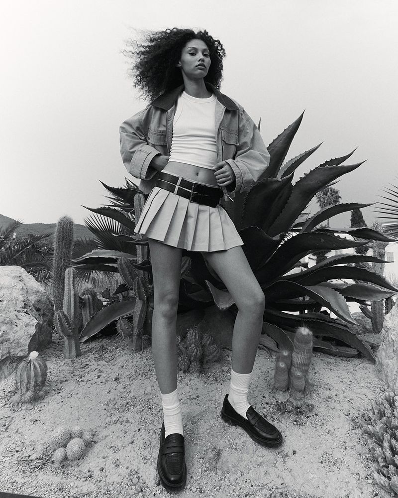 A young woman with curly hair is posing in a pleated mini skirt, jacket, socks and loafers, amidst desert flora in a black-and-white fashion shot.