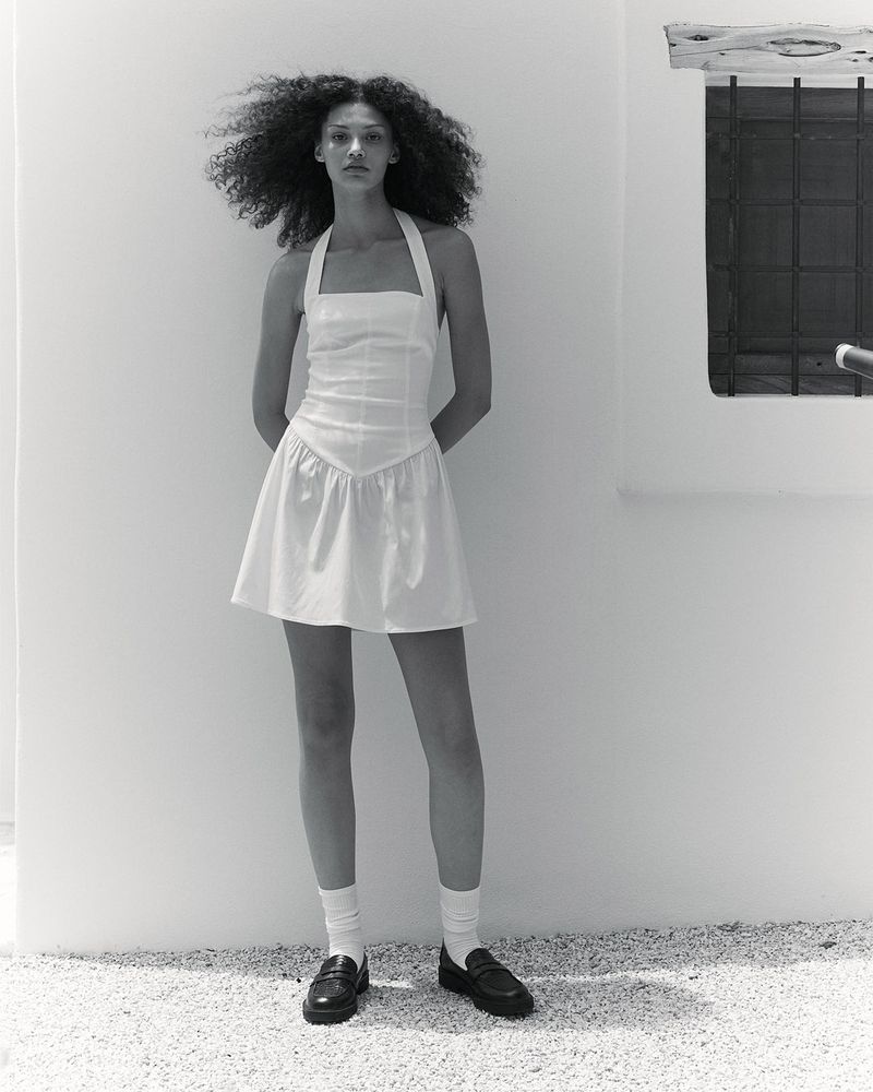 Model wearing a white halterneck mini dress, white socks, and black loafers standing against a sunlit exterior white wall.
