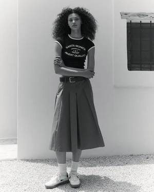Enya Davis standing in a black graphic ringer t-shirt, pleated midi skirt, and white sneakers against a whitewashed wall.