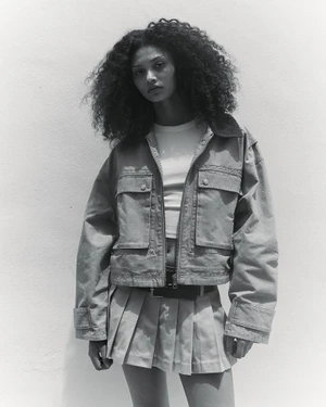 Model Enya Davis wearing a cropped utility jacket and pleated mini skirt in a black and white portrait against a light backdrop.