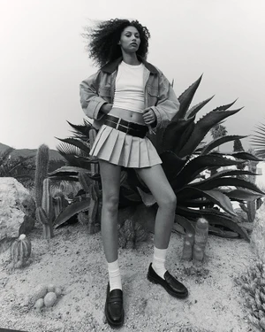 Model Enya Davis poses outdoors in a pleated mini skirt, cropped top, jacket, and loafers against agave plants.
