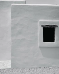 Black and white architectural detail of a white textured stucco wall with a small recessed square window.