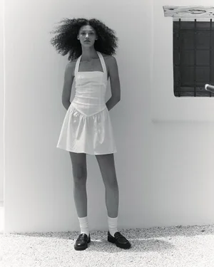 Model wearing a white halterneck mini dress, white socks, and black loafers standing against a sunlit exterior white wall.