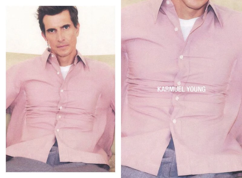 Portrait of an adult male wearing a pink button-down shirt and grey pants, casually posing indoors with a neutral background.