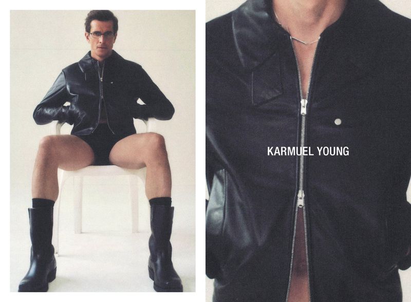 Man sits on a white chair in a photo studio wearing glasses, a leather jacket, boots, and black underwear against a white background.