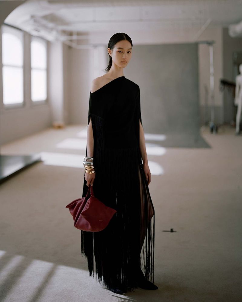 Model wearing a black fringe dress holding a red handbag in a loft studio space for Altuzarra