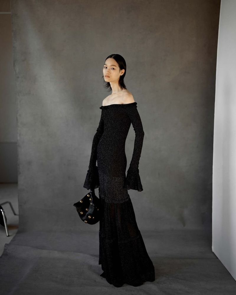 Model in long black off-the-shoulder dress holding a black handbag against a grey textured studio backdrop