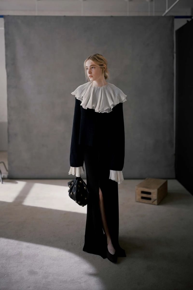 Elegant woman in long black dress with white ruffle collar and black purse, posing against gray studio backdrop with natural light.