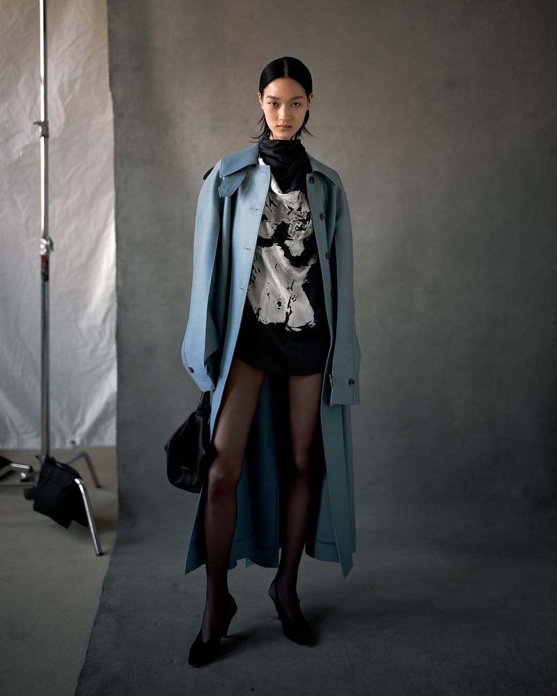 Asian woman poses in a studio, wearing a light-blue trench coat, black top, and heels against a gray backdrop with soft lighting.