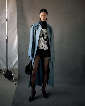 Asian woman poses in a studio, wearing a light-blue trench coat, black top, and heels against a gray backdrop with soft lighting.