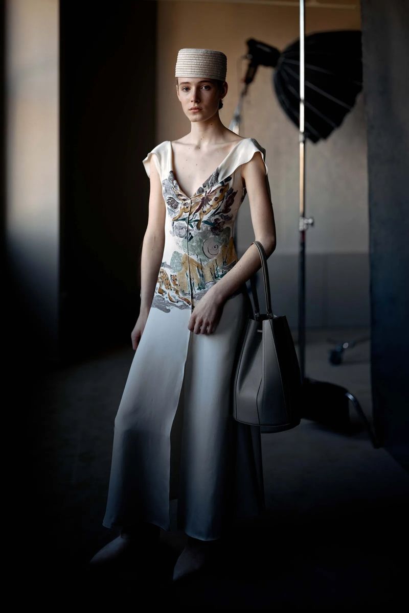 Model wearing a patterned dress and pillbox hat holding a bucket bag in a studio setting with lighting equipment