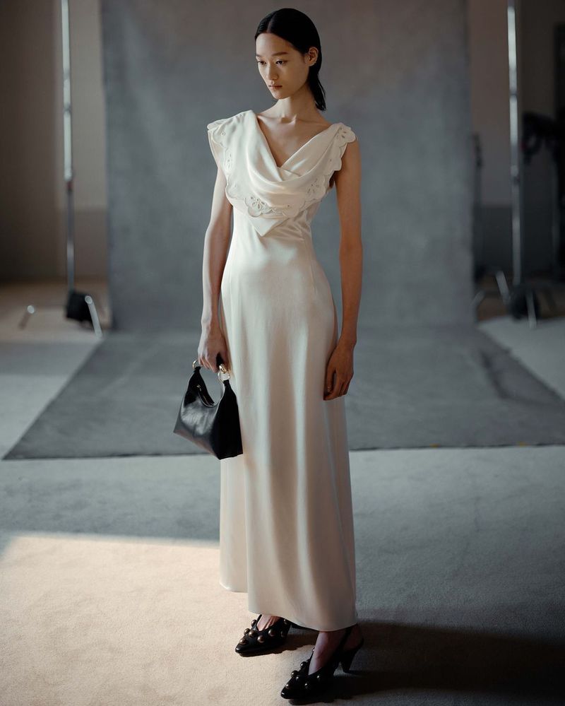 An Asian woman poses in a bright studio wearing a white dress and black heels, holding a black handbag, creating a fashion editorial image.