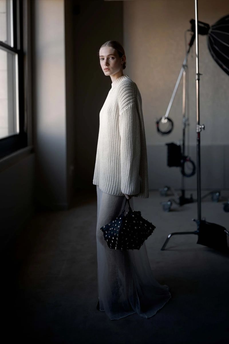 Model in cream ribbed knit sweater and sheer trousers holding a black handbag in a studio