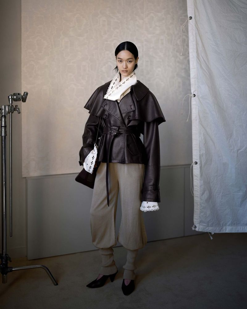 Fashion portrait of a woman wearing a brown leather jacket, beige pants, and black heels in a studio with neutral background.