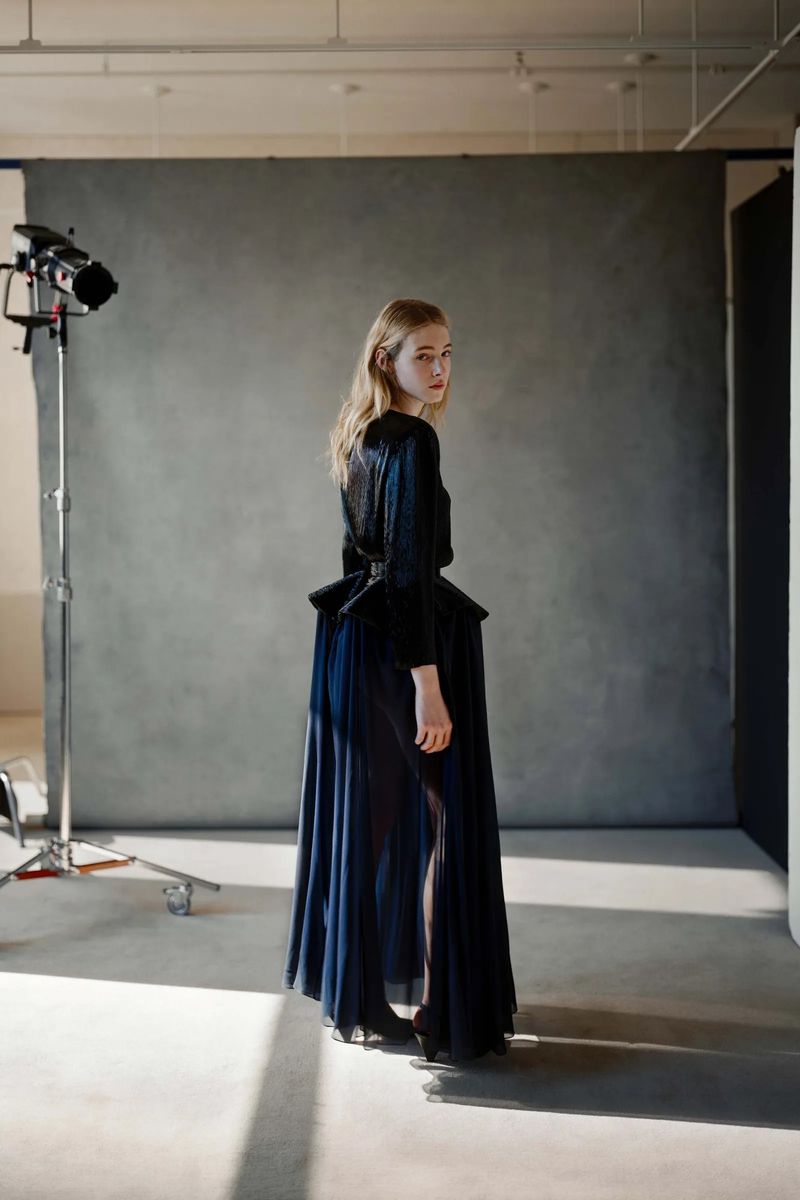 Fashion shot of a young, blonde woman in a dark dress with a full skirt in a photo studio, looking over her shoulder toward the camera.