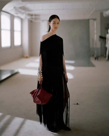 Model wearing a black fringe dress holding a red handbag in a loft studio space for Altuzarra