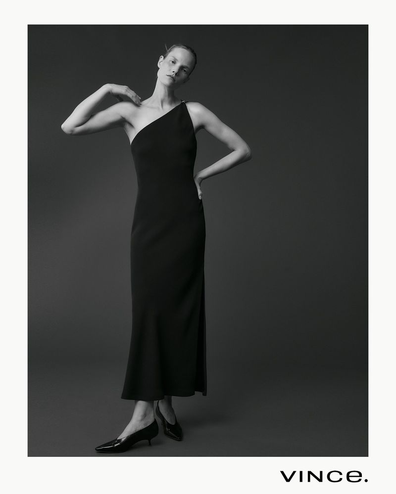 Model Suvi Riggs wearing a black one-shoulder maxi dress and low heels against a dark studio background.