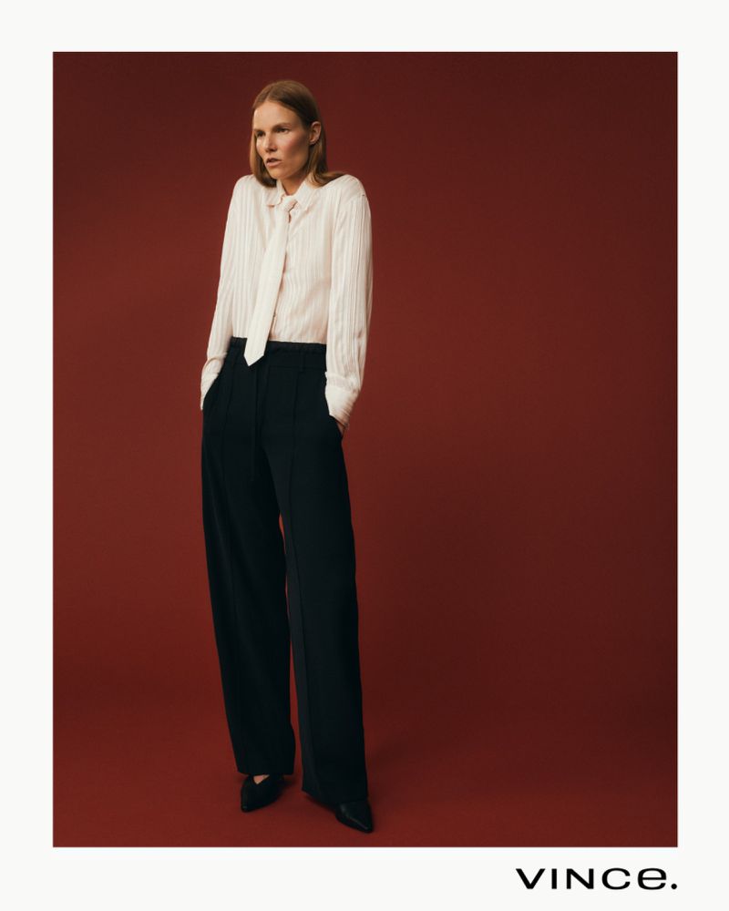 Studio shot of a woman wearing a white blouse with a neck tie and black trousers against a solid red background.