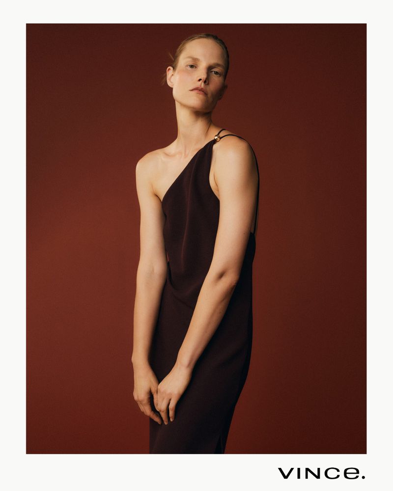 Elegant woman in a one-shoulder black dress posing against a maroon backdrop in a fashion editorial shot, showcasing minimalist style and design.