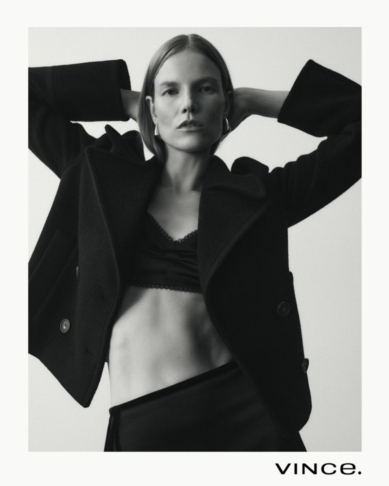 Black and white portrait of a female model in a black coat and lace top posing in a photo studio with soft lighting and neutral background.