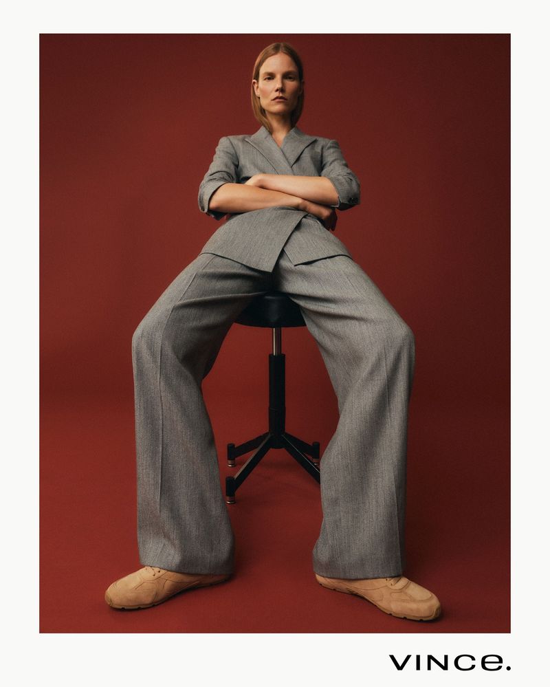 Fashion shot of an adult female in a grey suit sitting on a stool against a red backdrop with the brand name VINCE at the bottom.