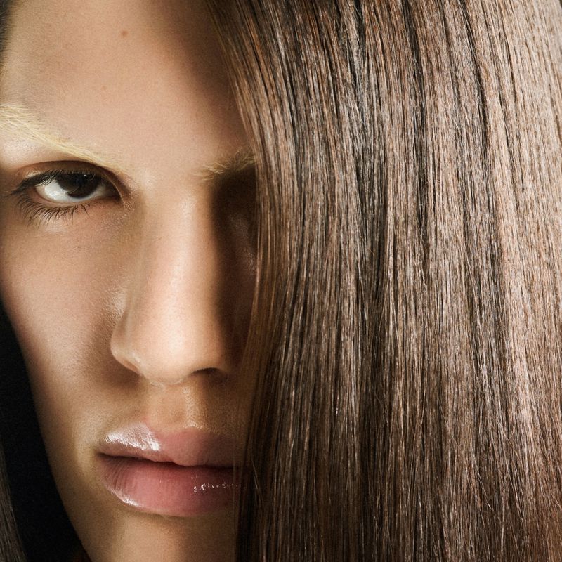 Close-up of a fashion model with straight brown hair partially covering the face, accented by light blonde eyebrows and glossy lips.