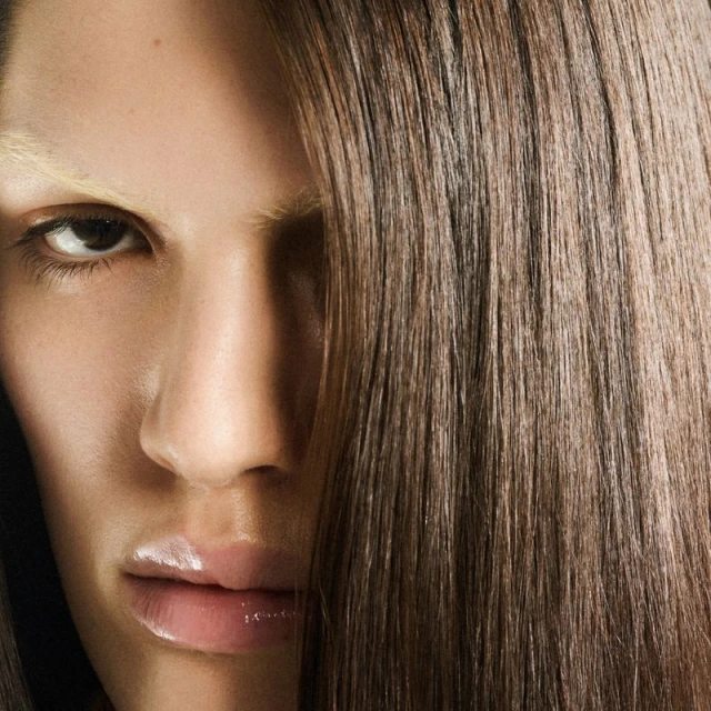 Close-up of a fashion model with straight brown hair partially covering the face, accented by light blonde eyebrows and glossy lips.
