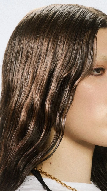 Close-up shot of a woman with shiny, dark brown wavy hair and minimal makeup wearing a gold chain necklace against a white background.