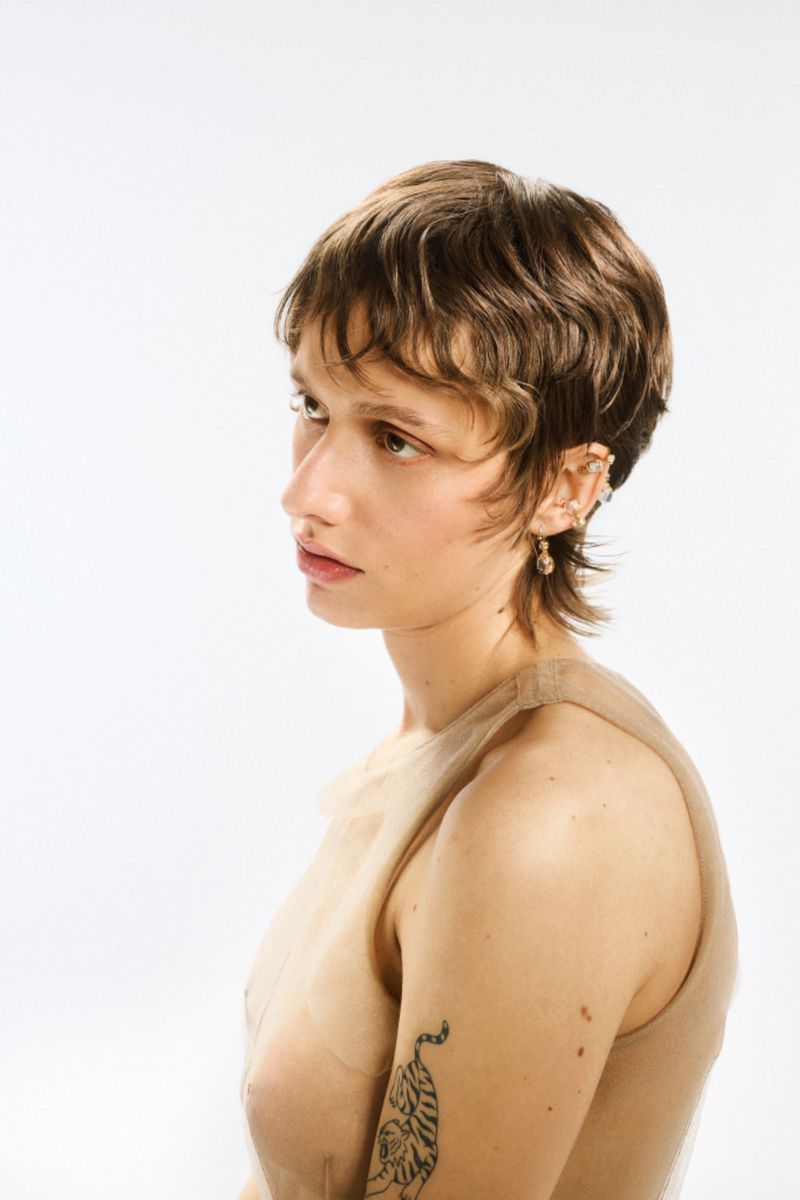 Portrait of a young adult woman with short brown hair, wearing a sheer beige top and earrings, in front of a bright white backdrop.