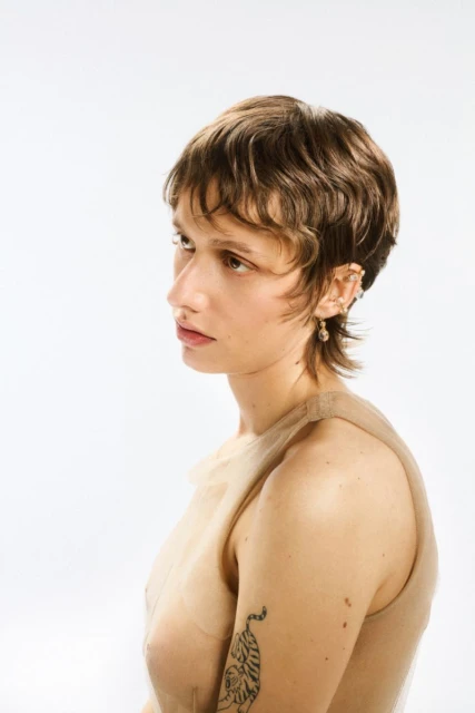 Portrait of a young adult woman with short brown hair, wearing a sheer beige top and earrings, in front of a bright white backdrop.