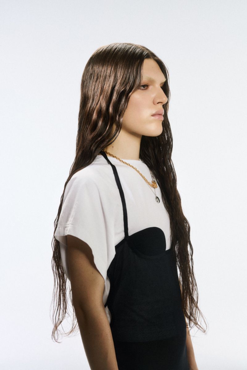 Woman with long brown hair wearing a black dress layered over a white t-shirt against a white backdrop in a fashion portrait.