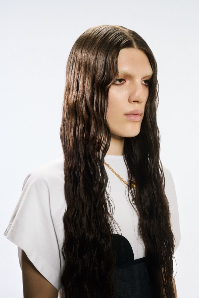 Portrait of a light-skinned adult with long, dark wavy hair, wearing a white t-shirt with a black detail, against a white background.