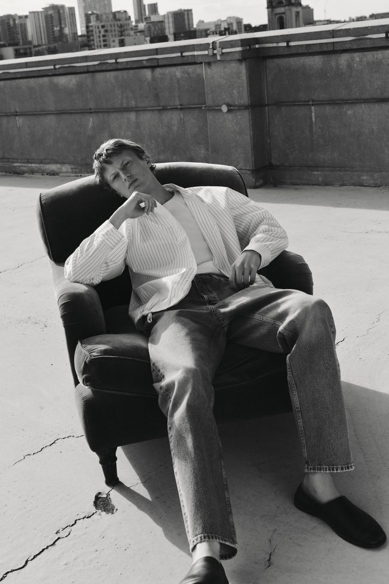 Black and white shot of a blonde adult male lounging in an armchair on a rooftop, wearing jeans and an unbuttoned pinstripe shirt.