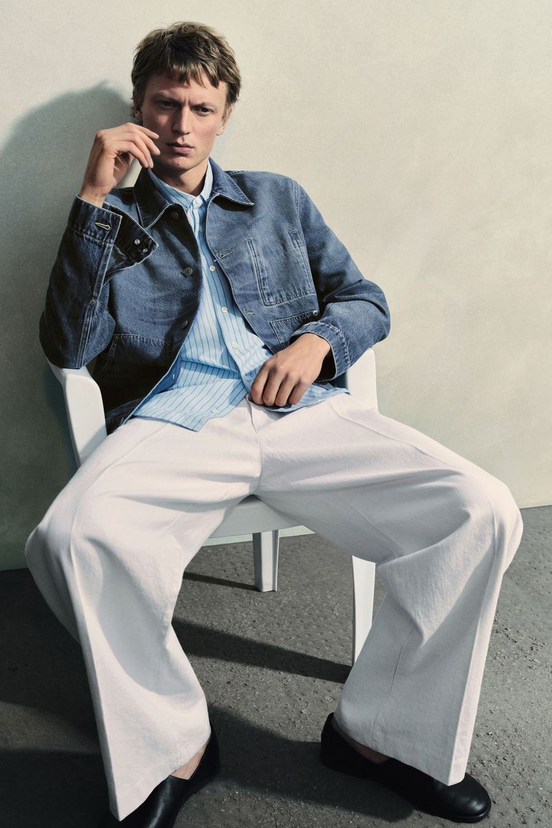 Man in a blue denim jacket, blue striped shirt, and white pants sits on a white chair in a studio setting.