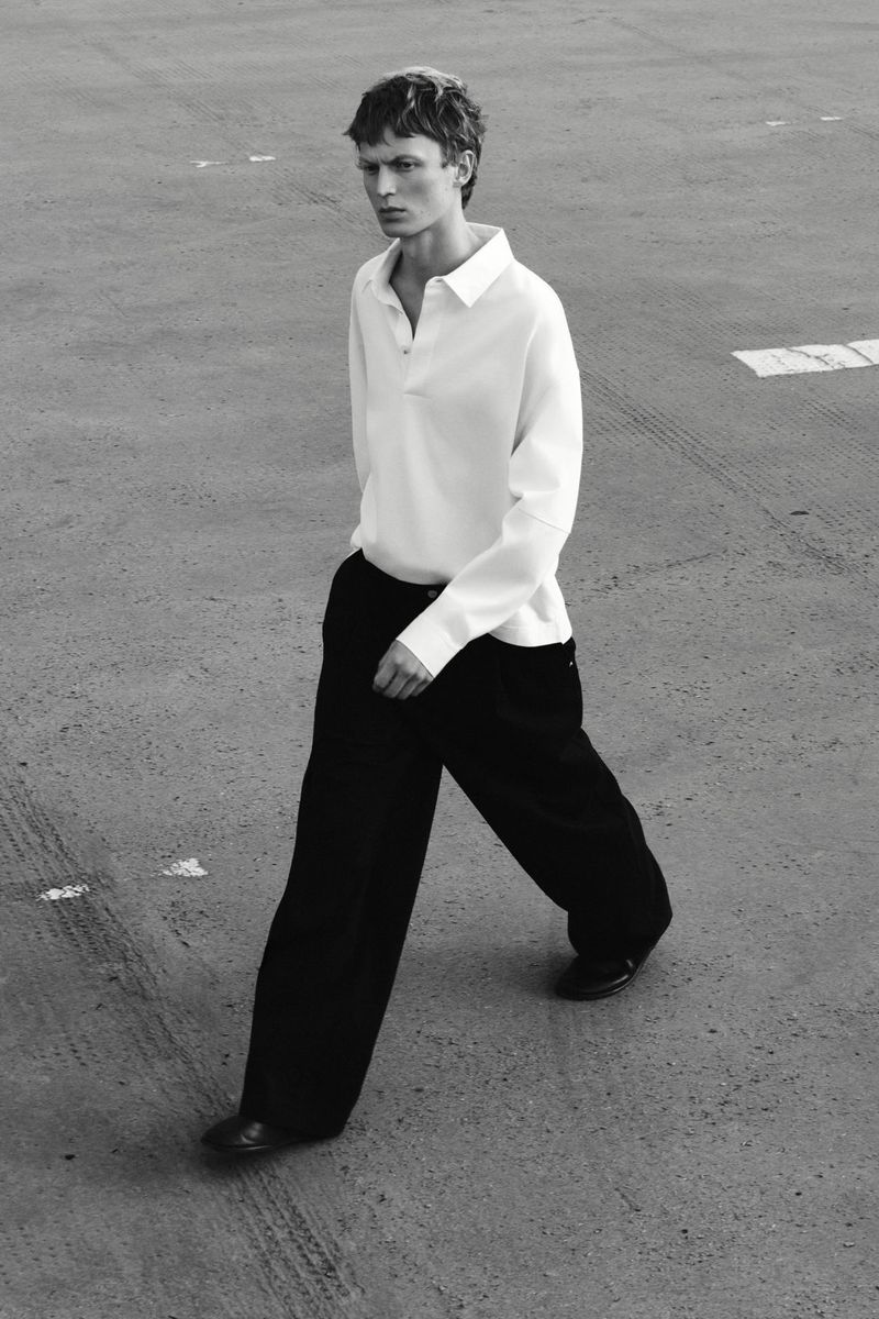 Monochrome full shot of a slender adult male walking outside wearing a white collared shirt, wide-legged black pants, and black shoes.