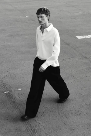 Monochrome full shot of a slender adult male walking outside wearing a white collared shirt, wide-legged black pants, and black shoes.