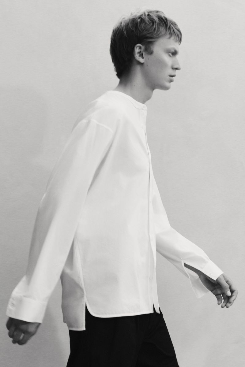 Studio shot of a young male model in a loose-fitting white shirt and black pants, walking against a light grey seamless backdrop.