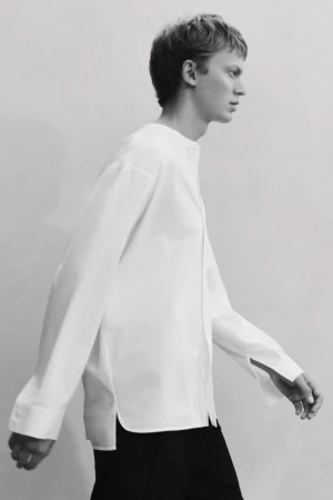 Studio shot of a young male model in a loose-fitting white shirt and black pants, walking against a light grey seamless backdrop.
