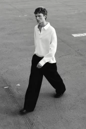 Model Jonas Glöer walking on an asphalt surface wearing a white long-sleeve polo shirt and wide-leg black trousers in black and white.