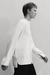 Model Jonas Glöer walks in profile wearing a white collarless long-sleeve shirt and black trousers against a plain studio background.