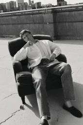 Model Jonas Glöer seated in an armchair on a rooftop wearing a striped jacket and jeans for COS Pre-Fall 2024.