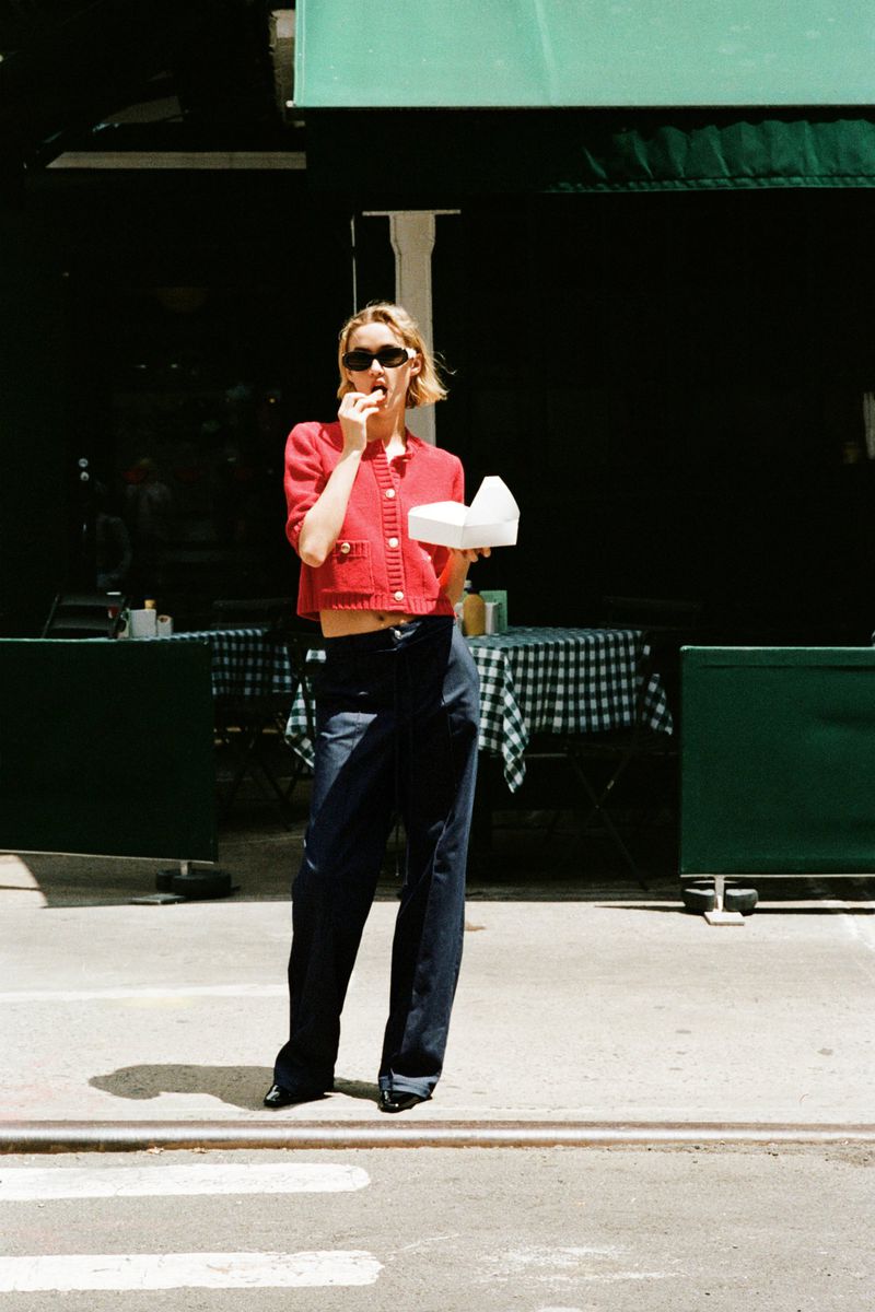 A woman in sunglasses and a red jacket is eating outside of a building with green trim, wearing dark pants and white sneakers.