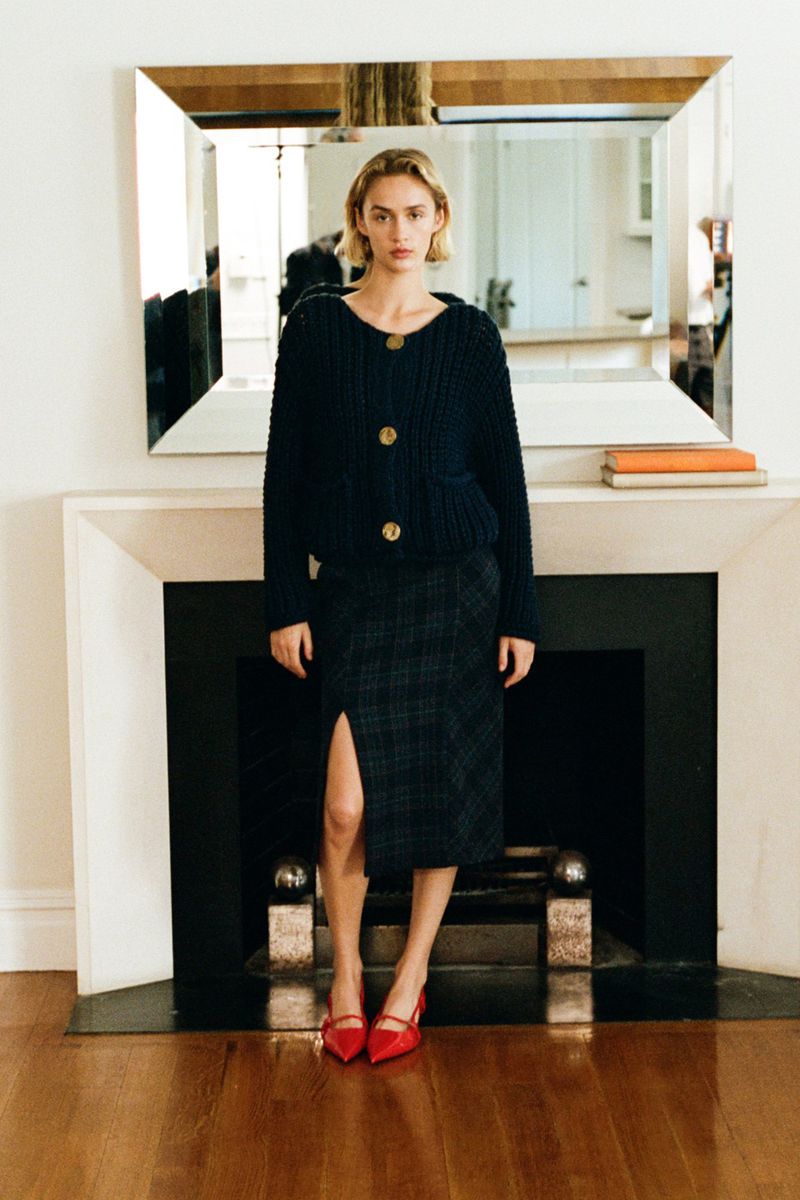 A fair-skinned woman with blonde hair poses indoors wearing a navy cardigan, plaid skirt with slit, and red heels in a portrait shot.