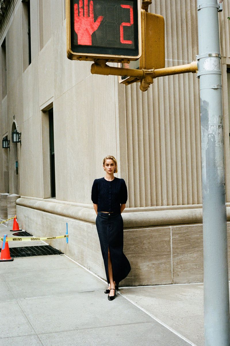 Woman in long skirt and sweater standing on city sidewalk with stop sign and building in background in an outdoor urban fashion shot.