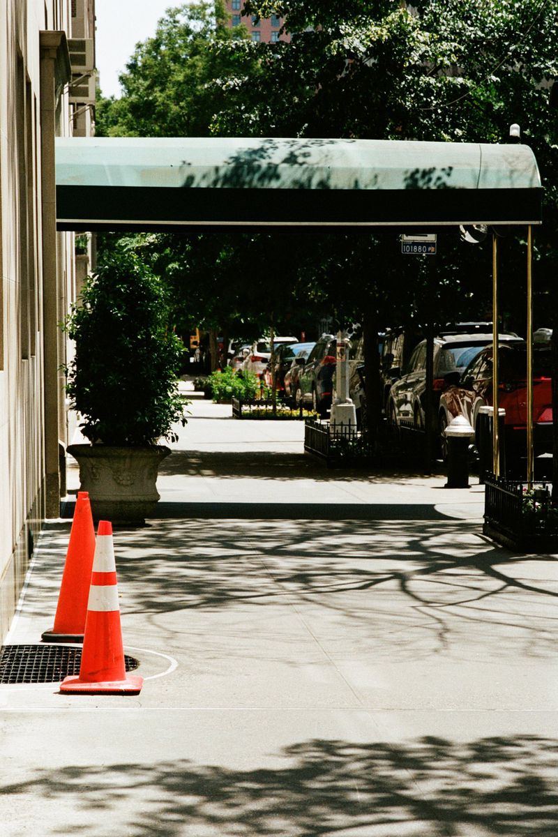 Outdoor shot of a sidewalk with two orange traffic cones and a building entrance covered by a green awning during daylight hours.