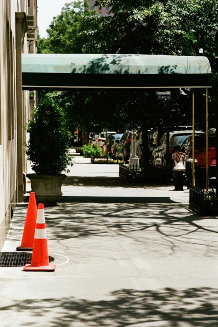 Outdoor shot of a sidewalk with two orange traffic cones and a building entrance covered by a green awning during daylight hours.