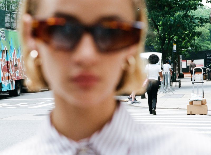 Close-up of a young woman with sunglasses and a striped shirt. She is walking on a city street with other pedestrians and vehicles.